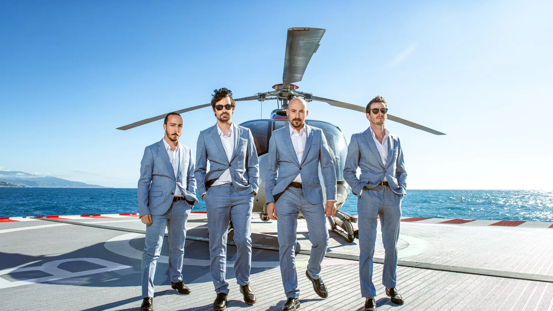 The band in light suits stepping off a helicopter on a seaside heliport
