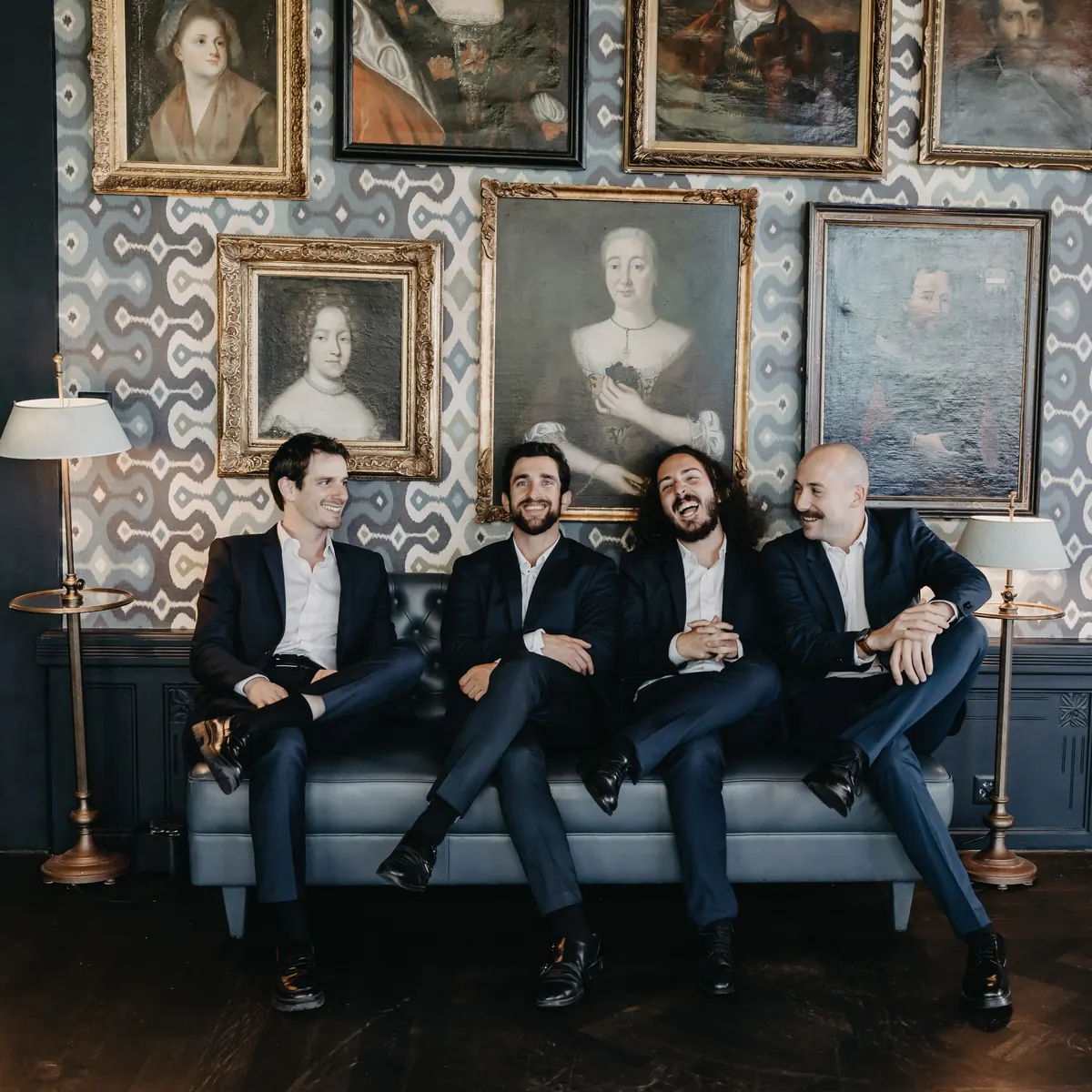 The band seated on a sofa under a wall of portraits, in dark suits