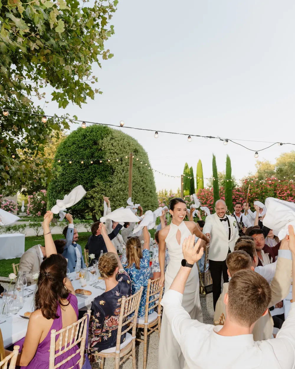 A long dinner table under the trees with guests raising their glasses